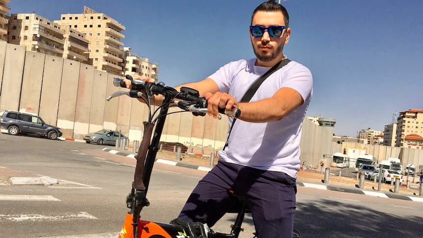 Palestinian nurse Firas, 26, rides a bike close to a tall concrete wall at Shuafat camp, West Bank.