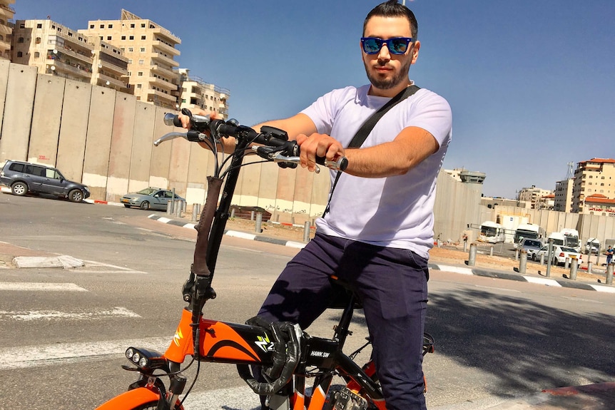 Palestinian nurse Firas, 26, rides a bike close to a tall concrete wall at Shuafat camp, West Bank.