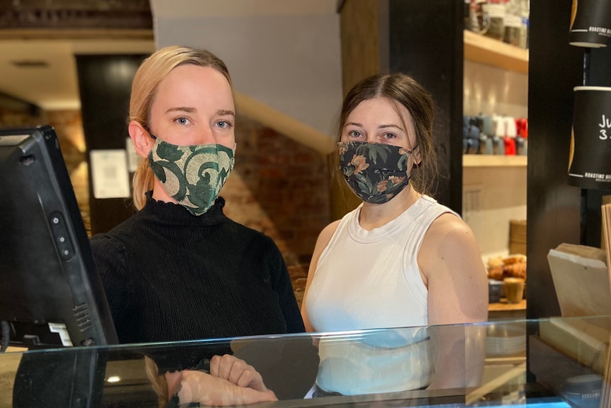 Paige and Rebecca stand together, with masks on, behind a cafe bench filled with focaccias and pastries.