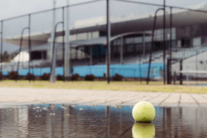 A tennis ball sits in a puddle in front of a tennis court on a cloudy day.