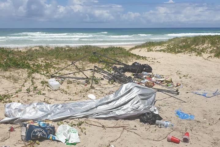 Beach strewn with a broken tent, bottles, and garbage bags left by campers.