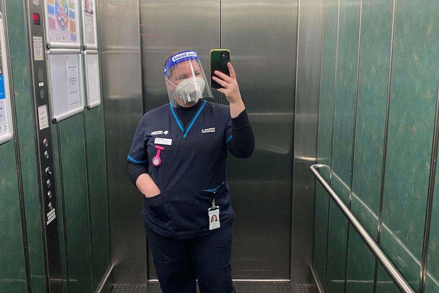 A woman standing in a lift with PPE and in scrubs.