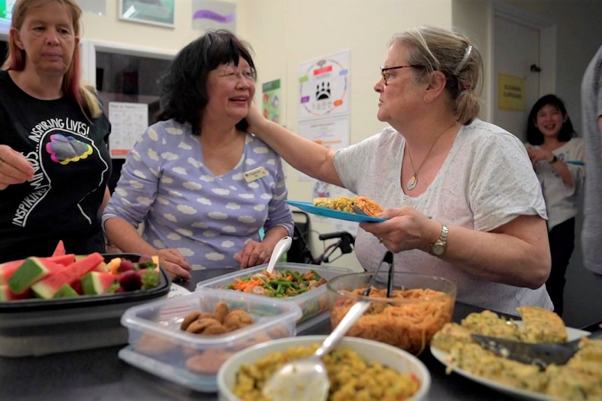 One woman reaches out to another in front of a table of food at a choir rehearsal.