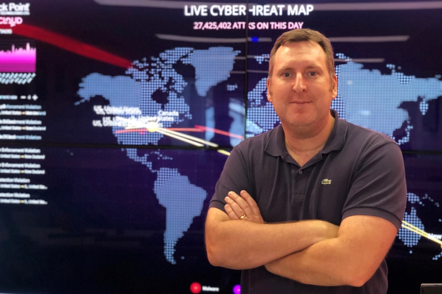 A man stands in front of a screen with a map of the world, his arms are crossed and he looks stern
