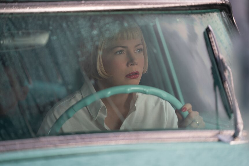 A white woman with a short, neat blonde bob sits at the wheel of a 50s style car in the rain and looks out.