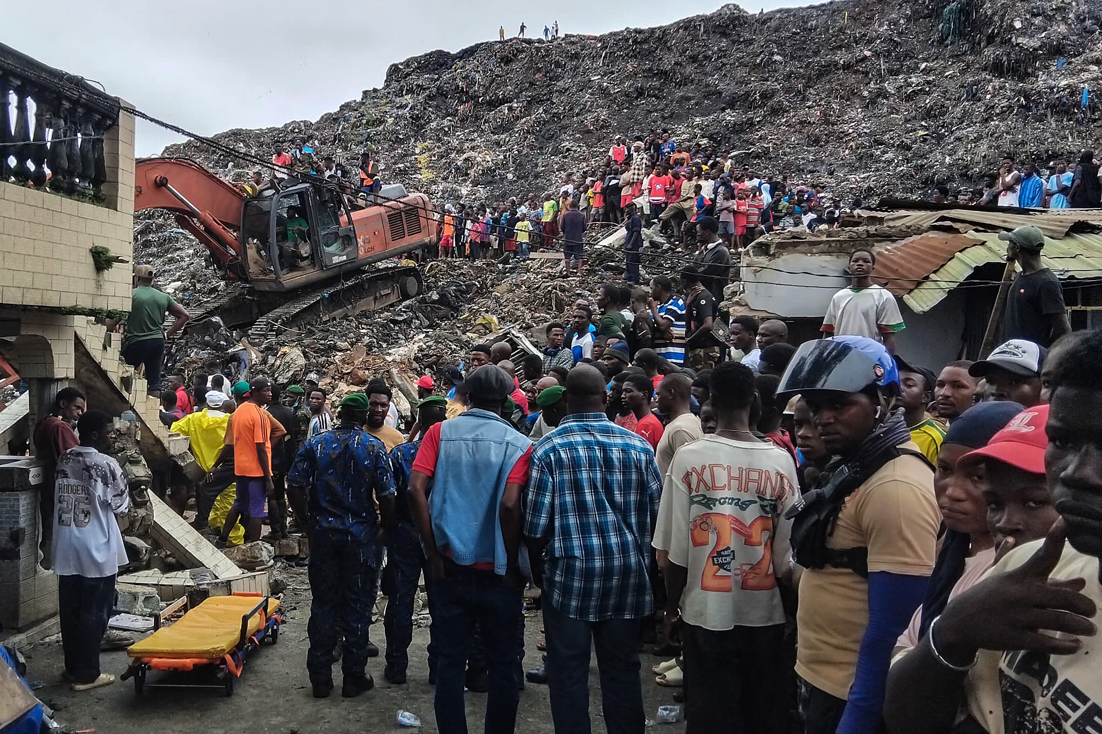 A group of people stand around an excavator on a pile of landfill.