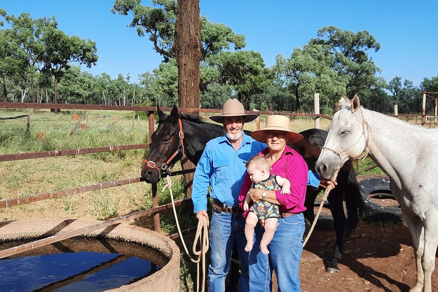 A man and a woman holding a baby stand in front of two horses next to a drinking trough in the outback.