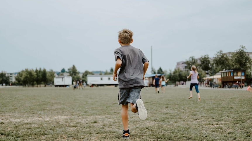 A boy seen from behind runs across a grassy field with other children playing in the distance