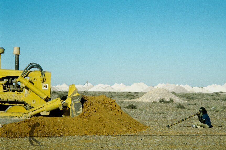 An Indigenous man sits playing didgeridoo opposite yellow mining vehicle, in the background are mounds of mullock or rock waste.