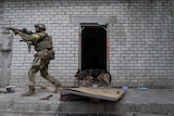 An armed soldier points a rifle forward as a patrol dog walks behind, past an open door.