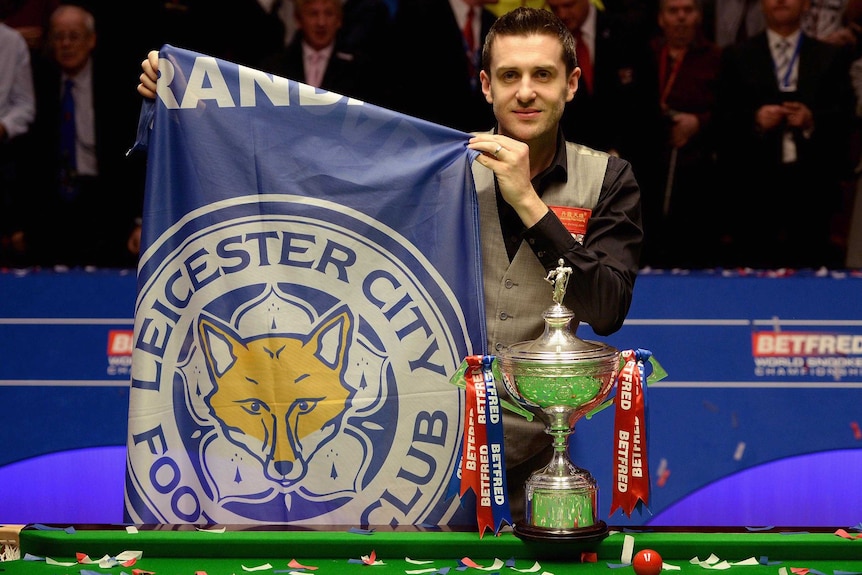 Mark Selby holds a Leicester City football club flag while standing next to the World Snooker Championship trophy.