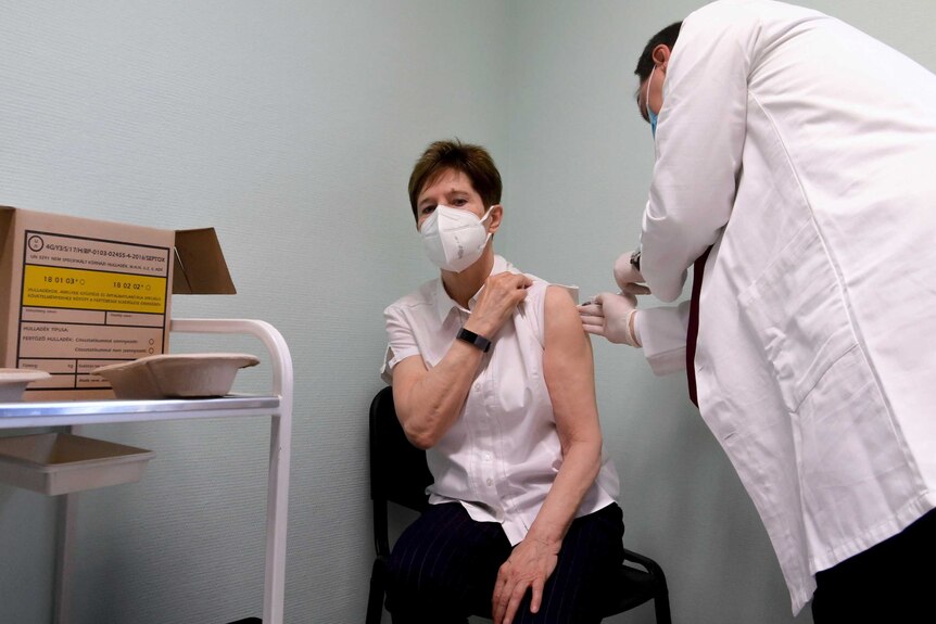 A middle-aged woman in face mask sits on chair in corner of room as white-coated doctor injects her.