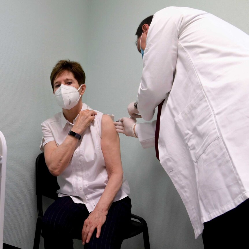 A middle-aged woman in face mask sits on chair in corner of room as white-coated doctor injects her.