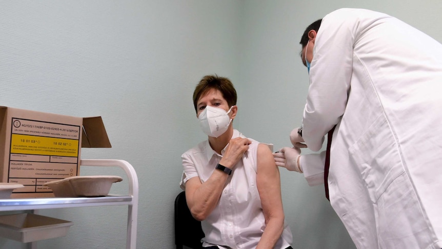 A middle-aged woman in face mask sits on chair in corner of room as white-coated doctor injects her.