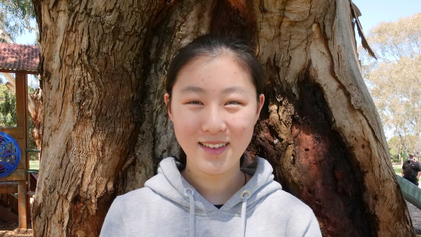 A young woman wearing a grey jumper stands in front of a tree, smiling.