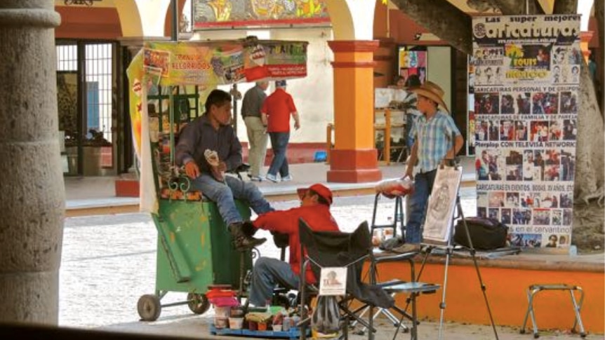 A man shining shoes in a square in Mexico.  His client sits on a green wheeled chair.