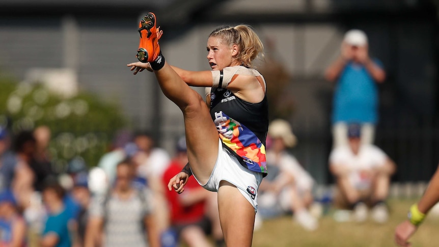 A woman wearing white football shorts, coloured singlet and orange football boots in mid-kick, with one leg high in the air.