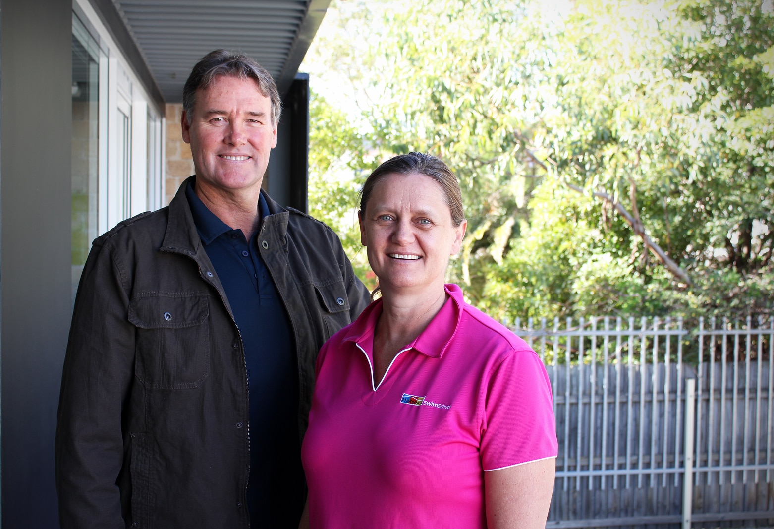 Ron wears a brown jacket and Susie wears a pink polo shirt outside, smiling at the camera.