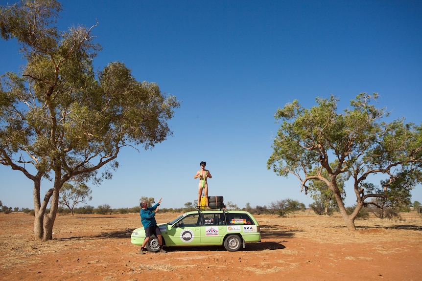 A man in a mankini stands on top of a brightly coloured station wagon in the desert. Another man points at him.