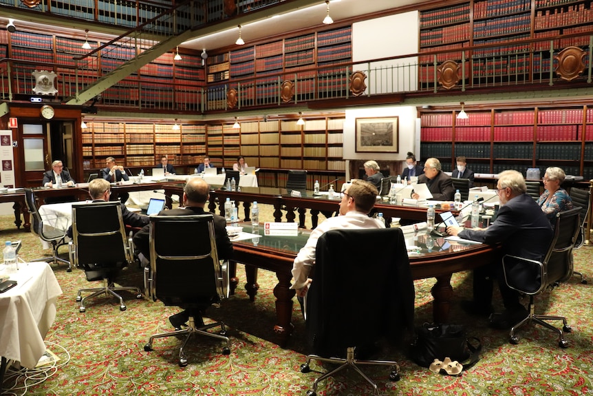 A dozen people sit around a large table facing one man who is being questioned.