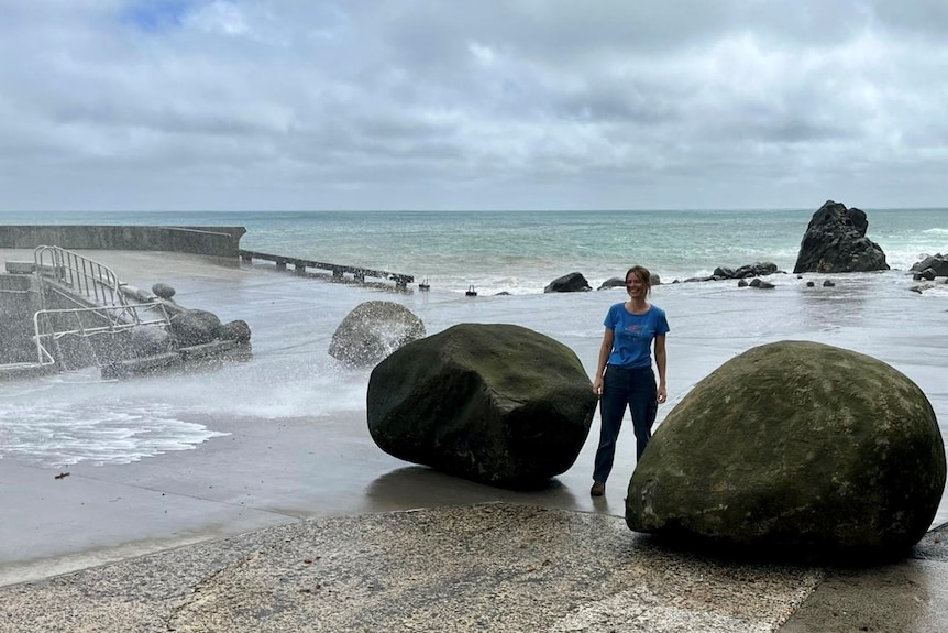 a woman standing between two boulders just a foot shorter than she is, wild seas behind her
