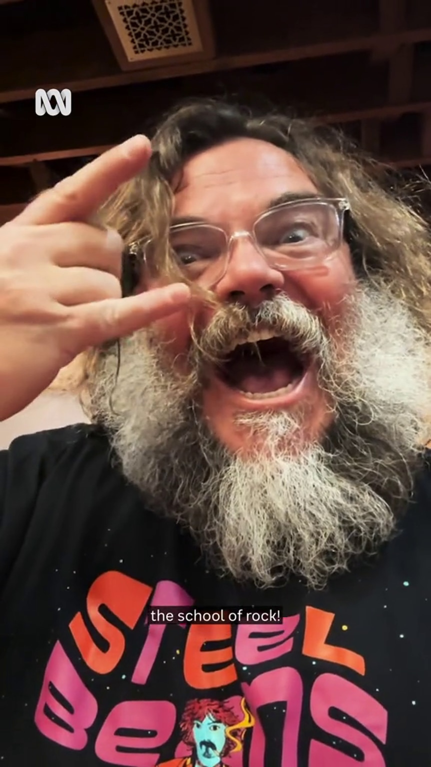 Jack Black, wearing glasses with a long beard, flashes the rock gesture with exaggerated facial expressions 