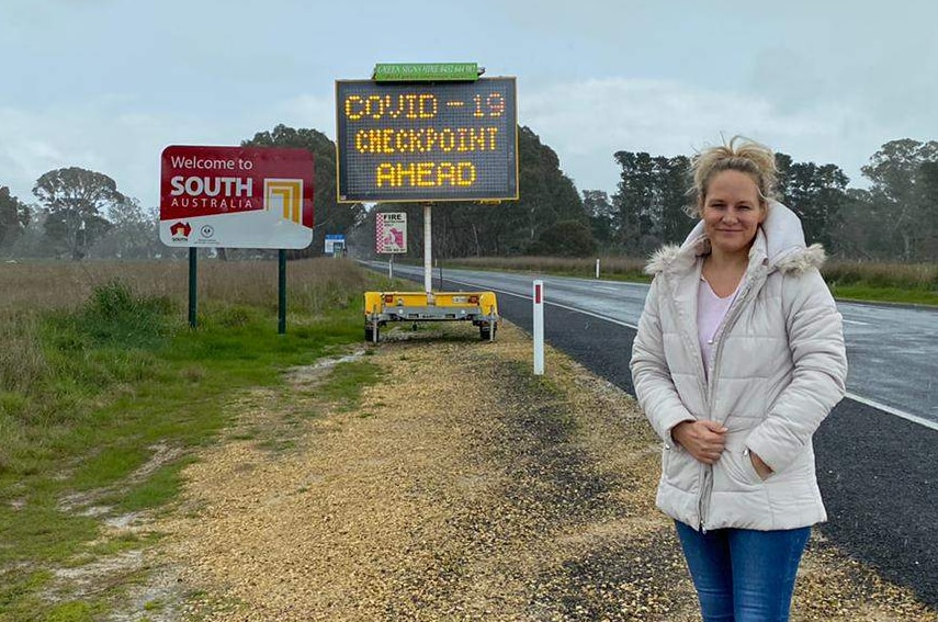 Paula Gust dressed in jeans and a coat stands in front of the "Welcome to South Australia" sign and COVID-19 checkpoint.