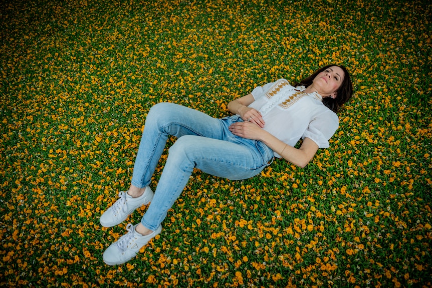 A woman with long black lying down on grass, surrounded by fallen leaves