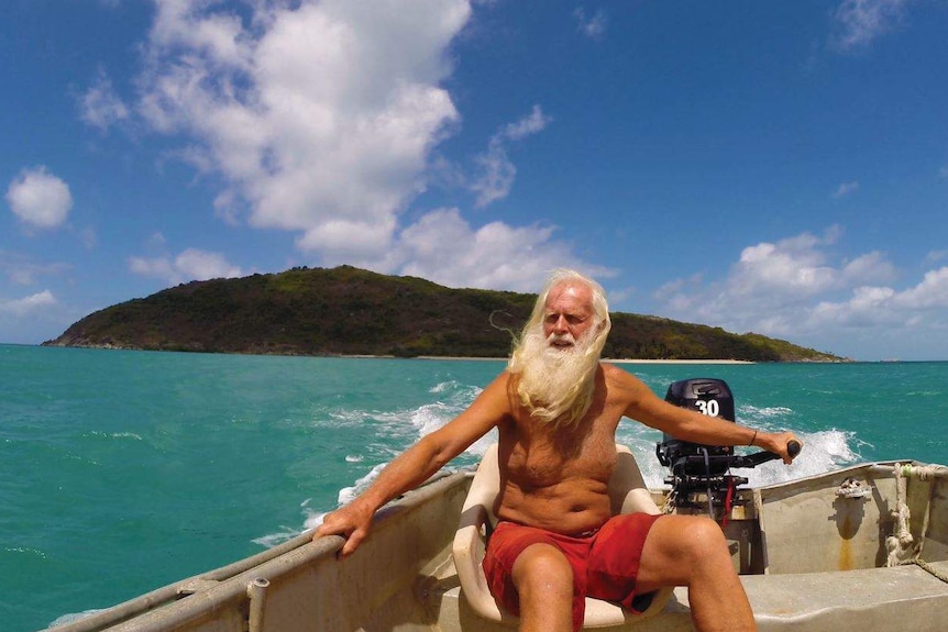 A man with long white hair and beard, and red shorts, sails a motorised tinny over turquoise water, with an island behind him.