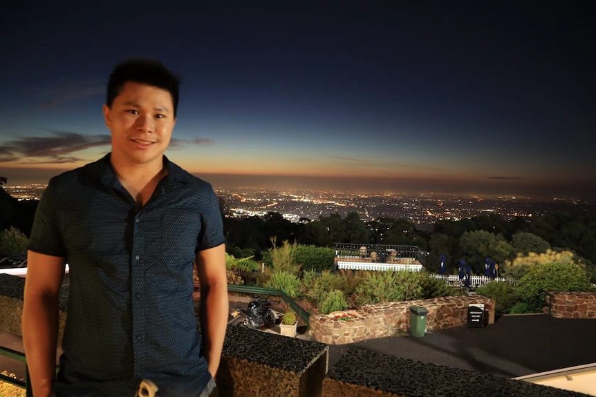 An Asian man standing on a balcony with a night view in the background