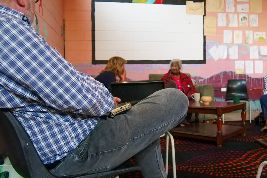 A man sits listening to a community discussion group in a colourful community hall.