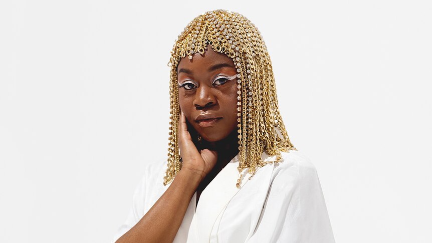 Sampa The Great wears a bejewelled, gold headpiece and stands against a stark white background