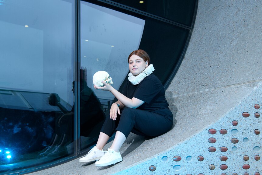 Young woman with auburn hair pulled back in a bun wears a white neck ruff, black clothes and holds a fibreglass skull.