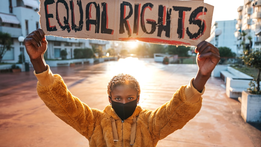 Young black woman with bleached hair and a mask on holding up a hand-made Equal Rights placard