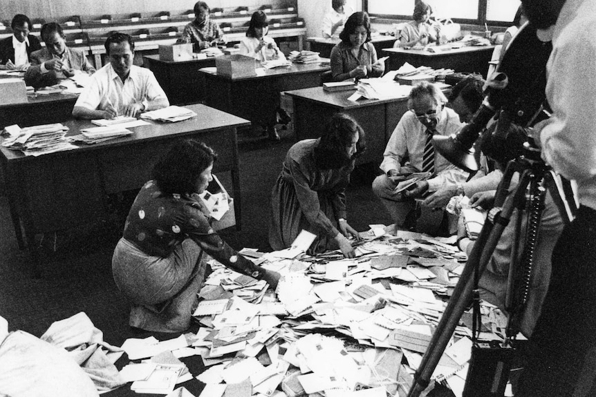 Piles of mail on the floor as people at desks open letters.