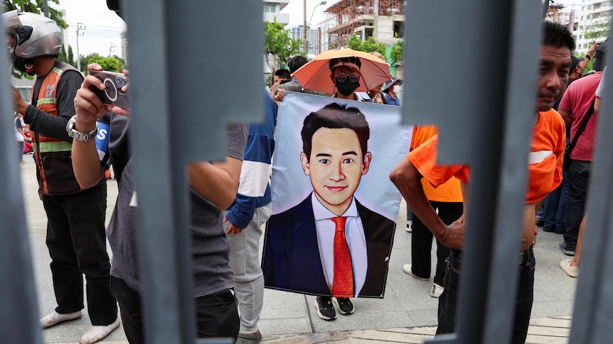 A man with his face covered and an umbrella holds a poster with a young Thai politician  