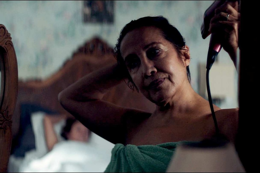 A woman in her sixties stares into a mirror, wrapped in a towel, drying her wet hair with a hairdryer.