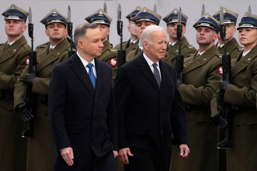 Two men in suits walk past a line of military personnel in dress uniform. 