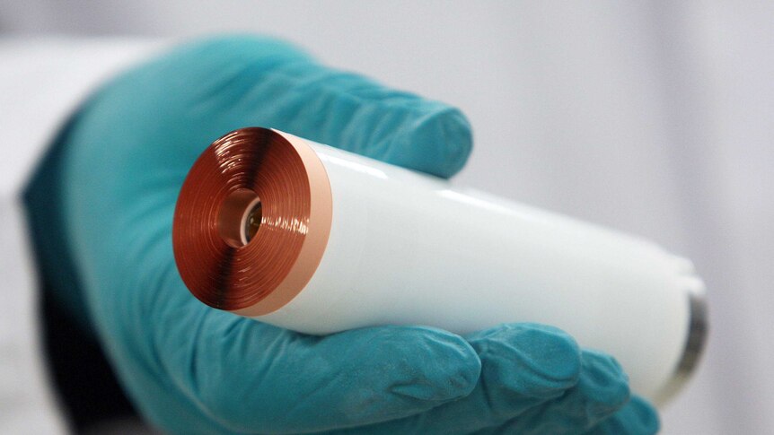 A gloved hand holds up a cylindrical lithium ion battery showing the rolls of lithium foil.