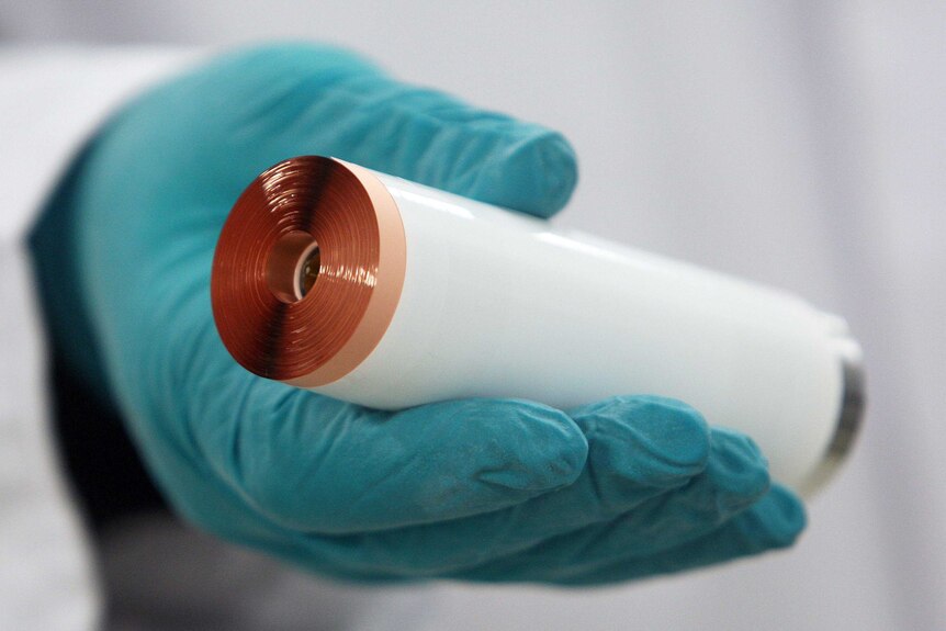 A gloved hand holds up a cylindrical lithium ion battery showing the rolls of lithium foil.