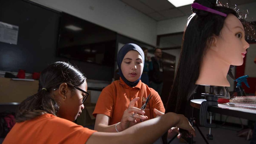 Two schoolgirls practice cutting the hair of a model dummy.