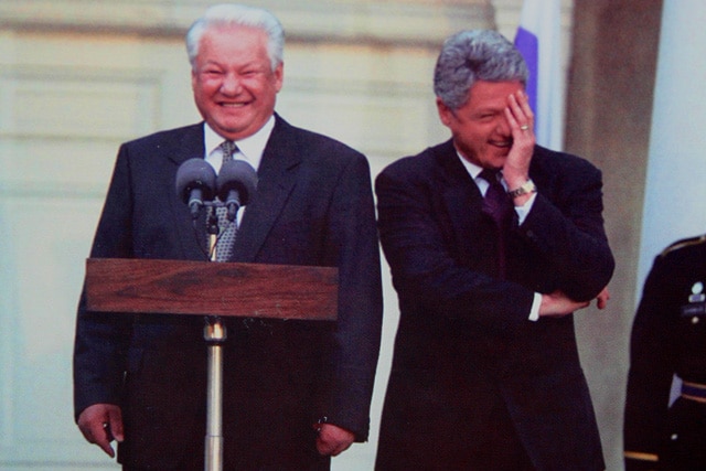 Two men in black suits stand behind a podium while laughing.  