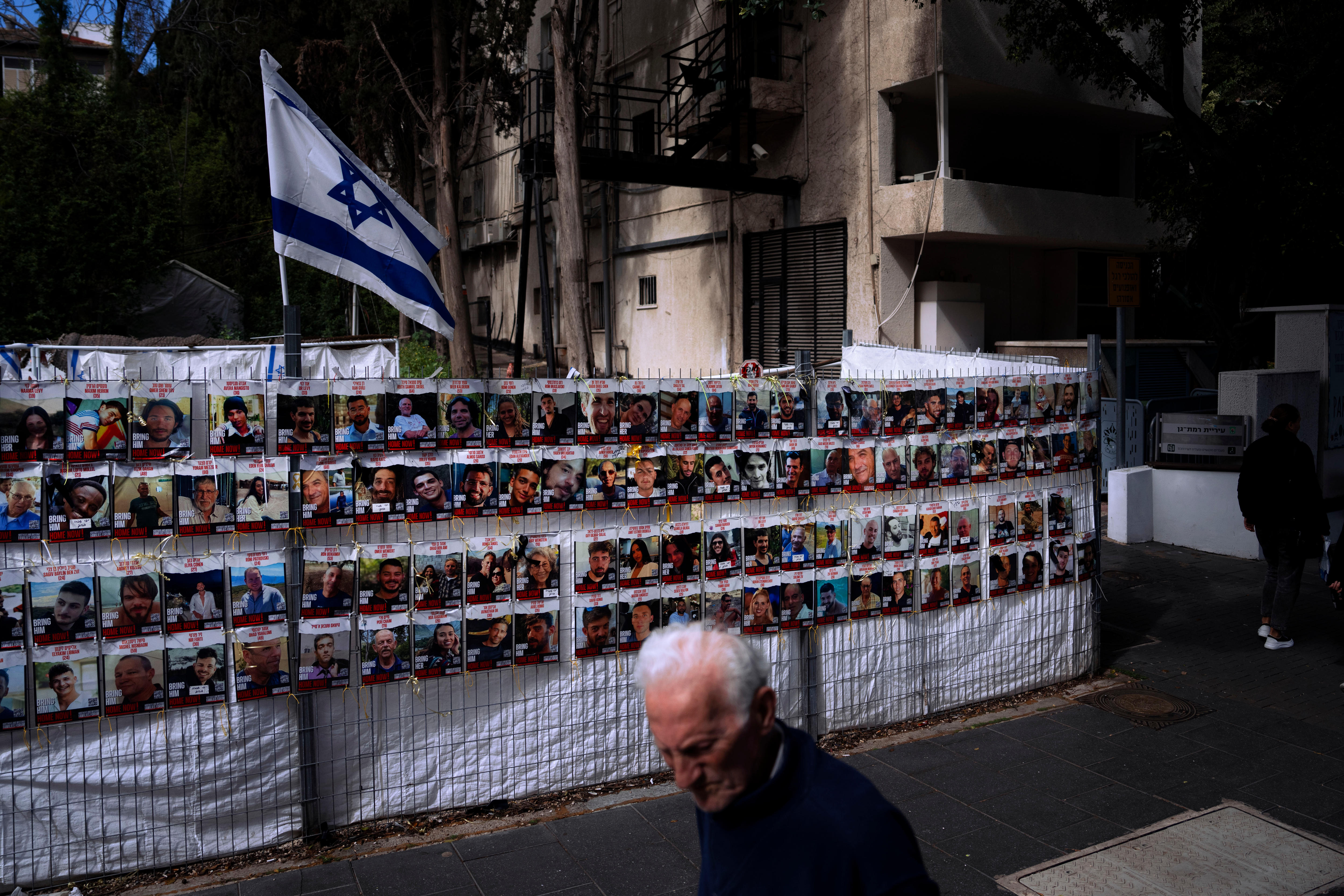 A man walks past a fence covered in photos of Israelis who are missing. A large Israeli flag flies above the fence.