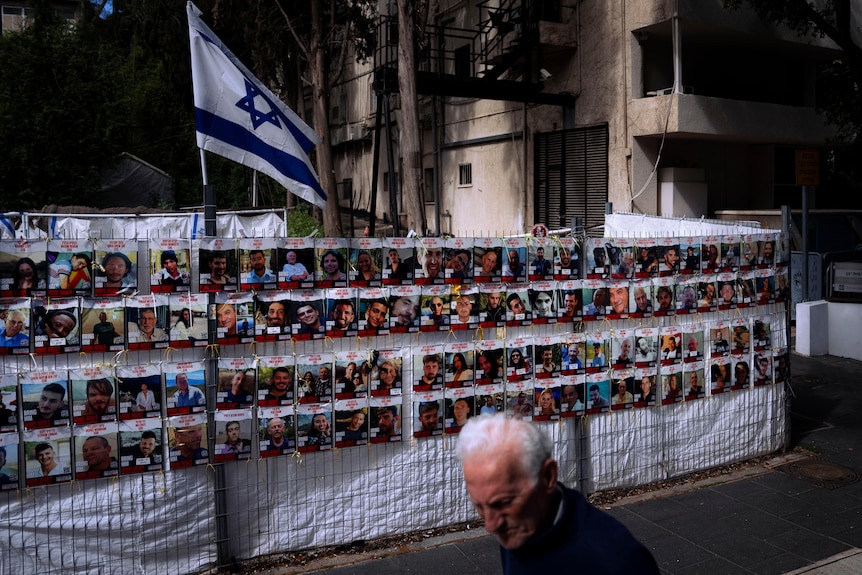 A man walks past a fence covered in photos of Israelis who are missing. A large Israeli flag flies above the fence.
