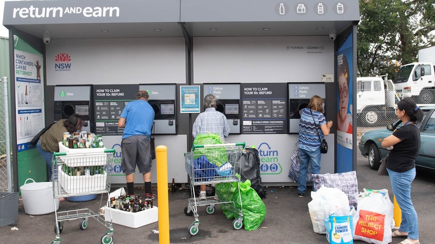 Four people empty bottles into a return and earn reverse vending machine in Marrickville in Sydney's west. Another lady waits.