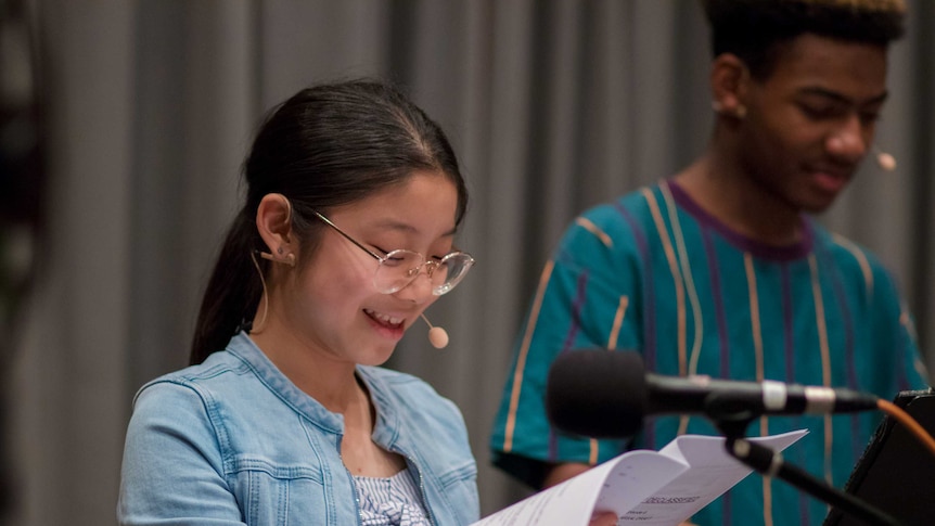 Teenage girl looking at script and talking into microphone with teenage boy in background.