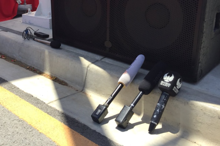 Microphones sit on the pavement leaning against a gutter, in front of a stack of speakers beside a stage.