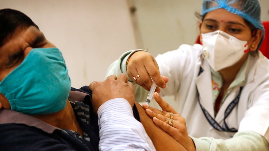 A health worker in PPE gives a vaccination to a man who winces while wearing a mask