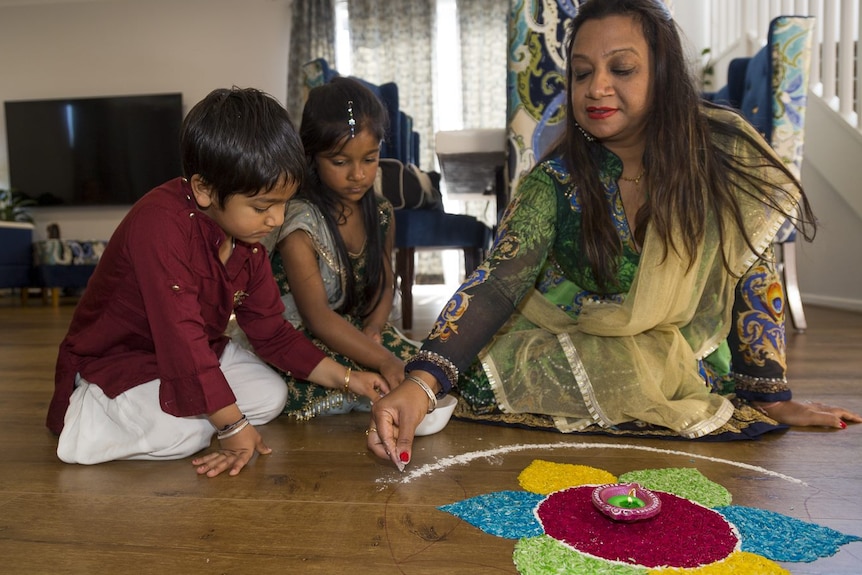 A woman is sitting on the floor, drawing a colourful pattern on the floor with a boy and a girl sitting besides her.