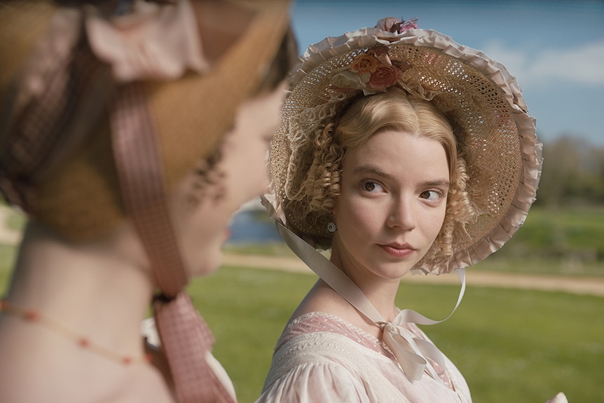 A blonde woman dressed in off-white Regency England period costume looks to the right at her walking companion on a sunny day.